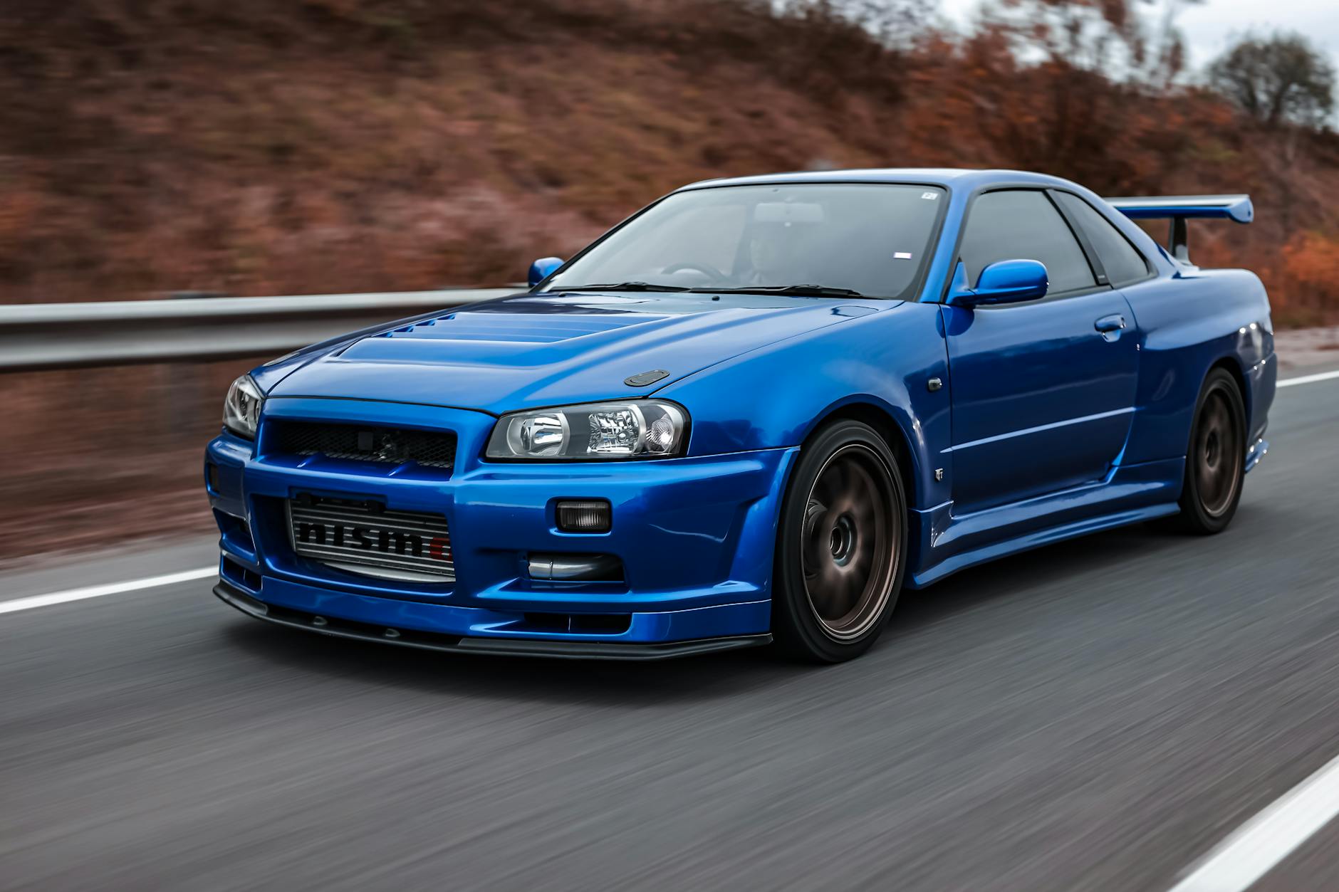 Ilustrasi: Close-up of a sleek blue Nissan Skyline GT-R driving on a scenic road.