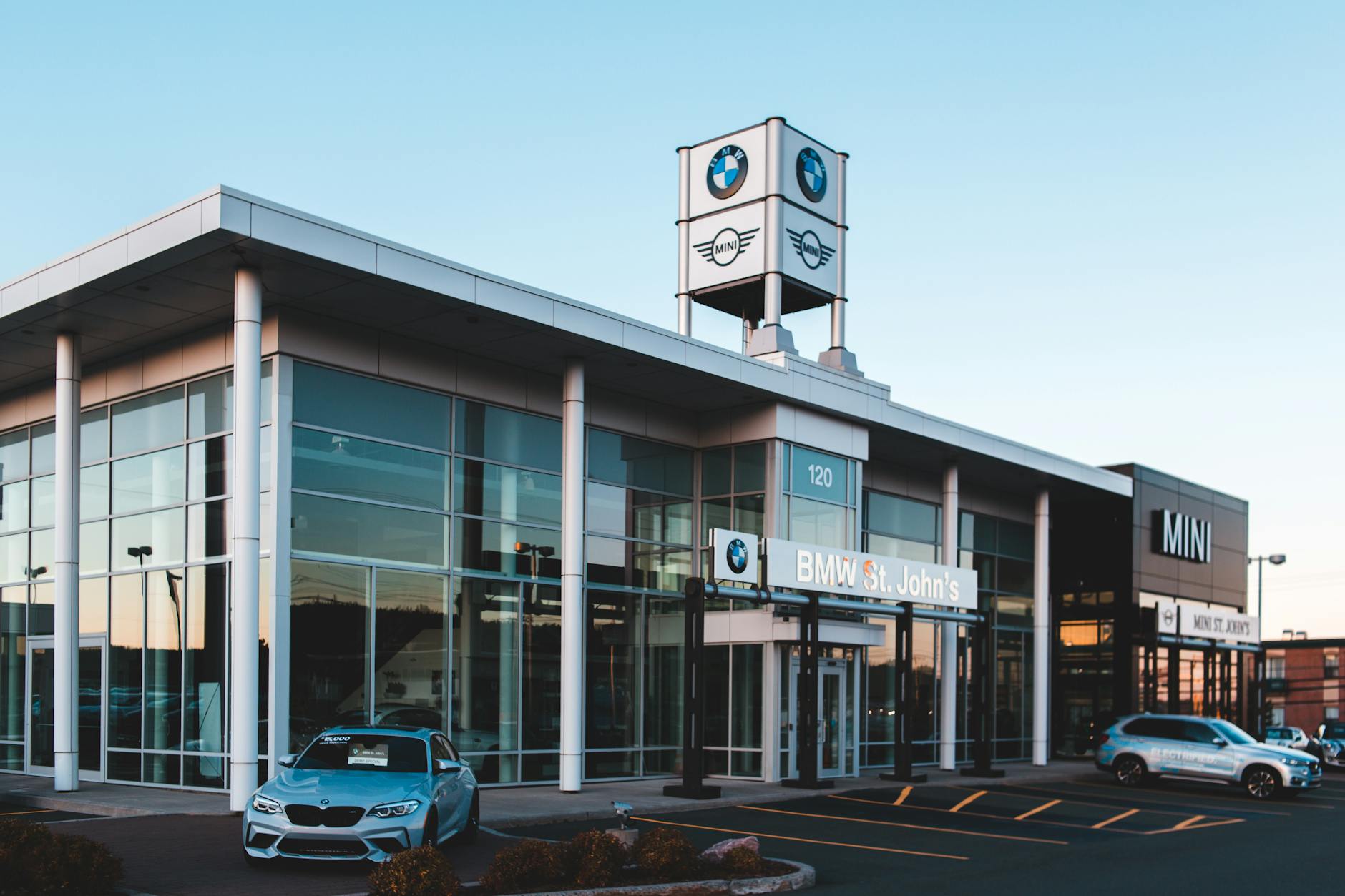Ilustrasi: Luxurious car dealership showcasing BMW and MINI cars with a modern glass exterior at dusk.