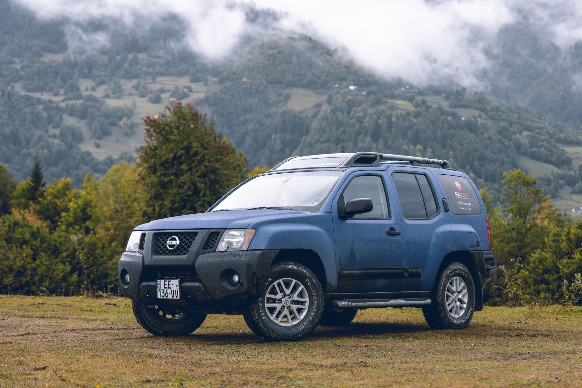 Ilustrasi: A rugged blue Nissan SUV parked against a picturesque mountain backdrop with lush greenery and misty clouds.