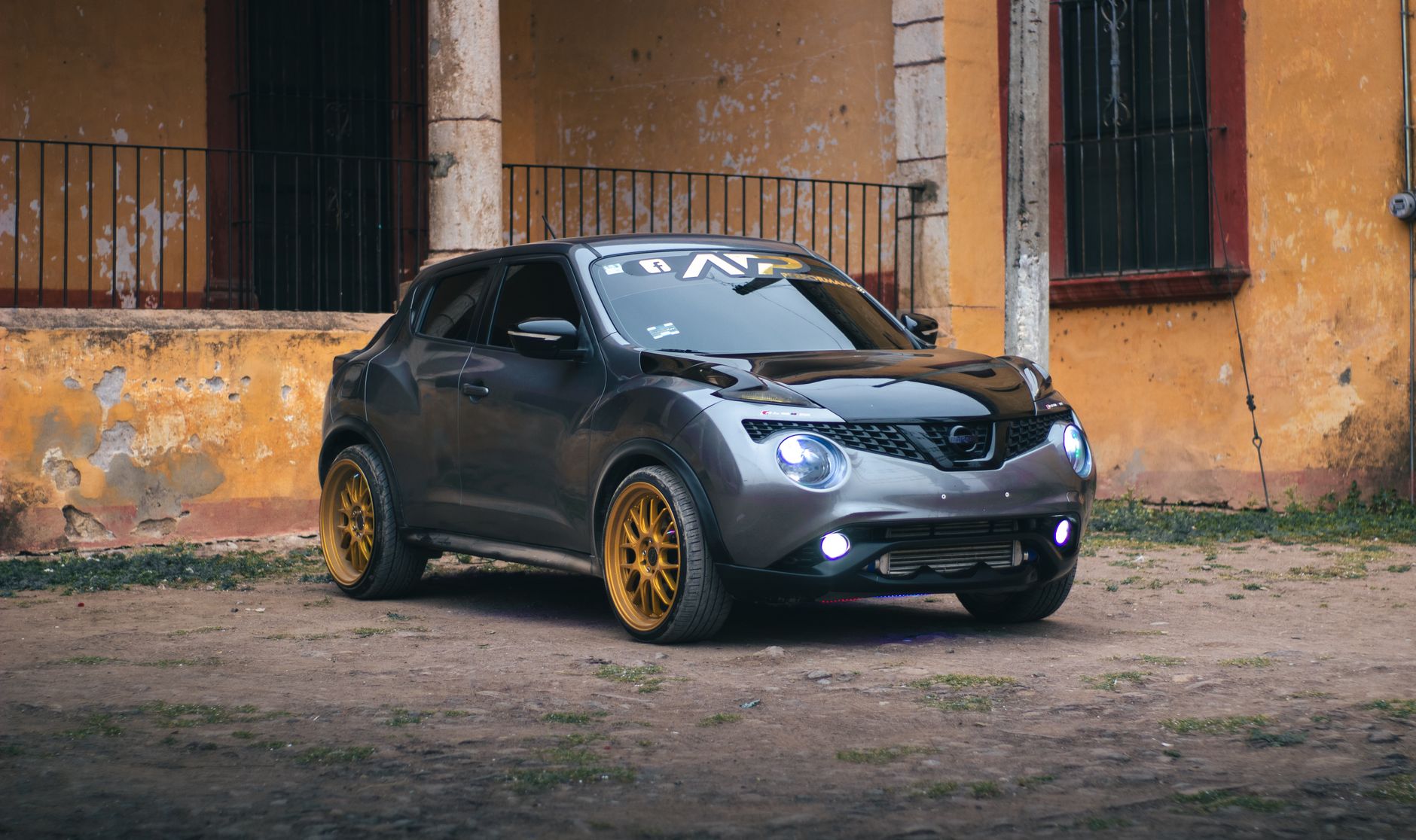 Ilustrasi: A sleek gray Nissan Juke with golden rims parked near a rustic building exterior.